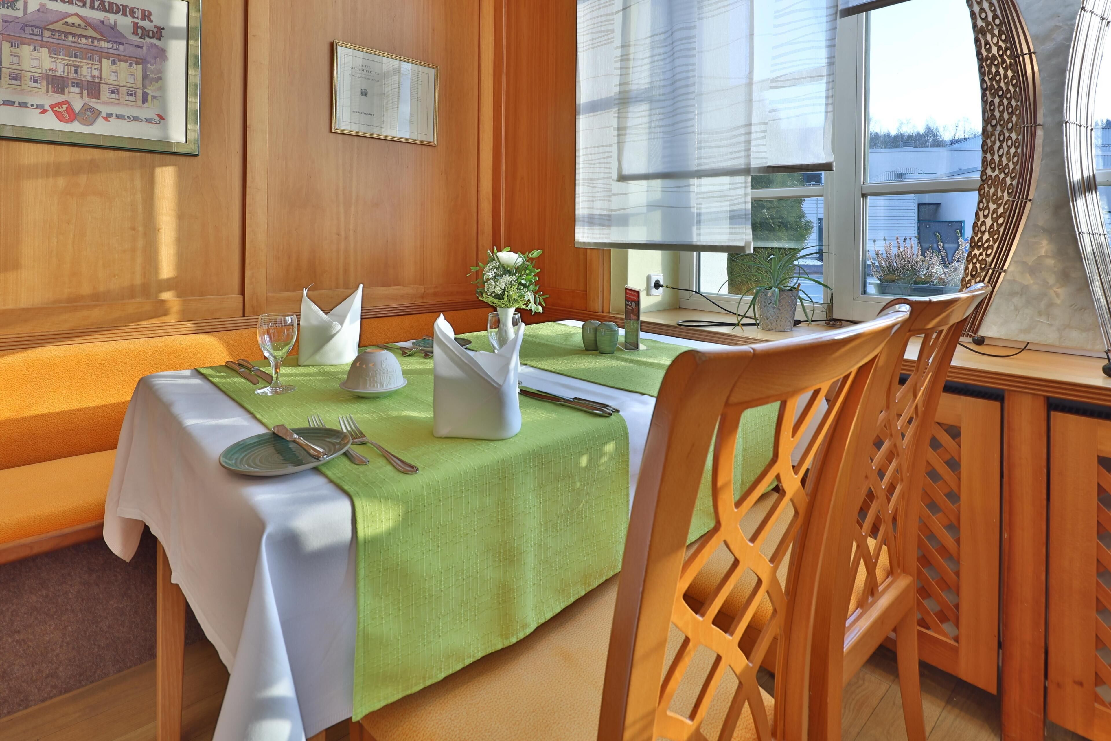 Un coin repas avec une table dressée pour deux personnes, un vase rempli de fleurs et une fenêtre donnant sur des immeubles.