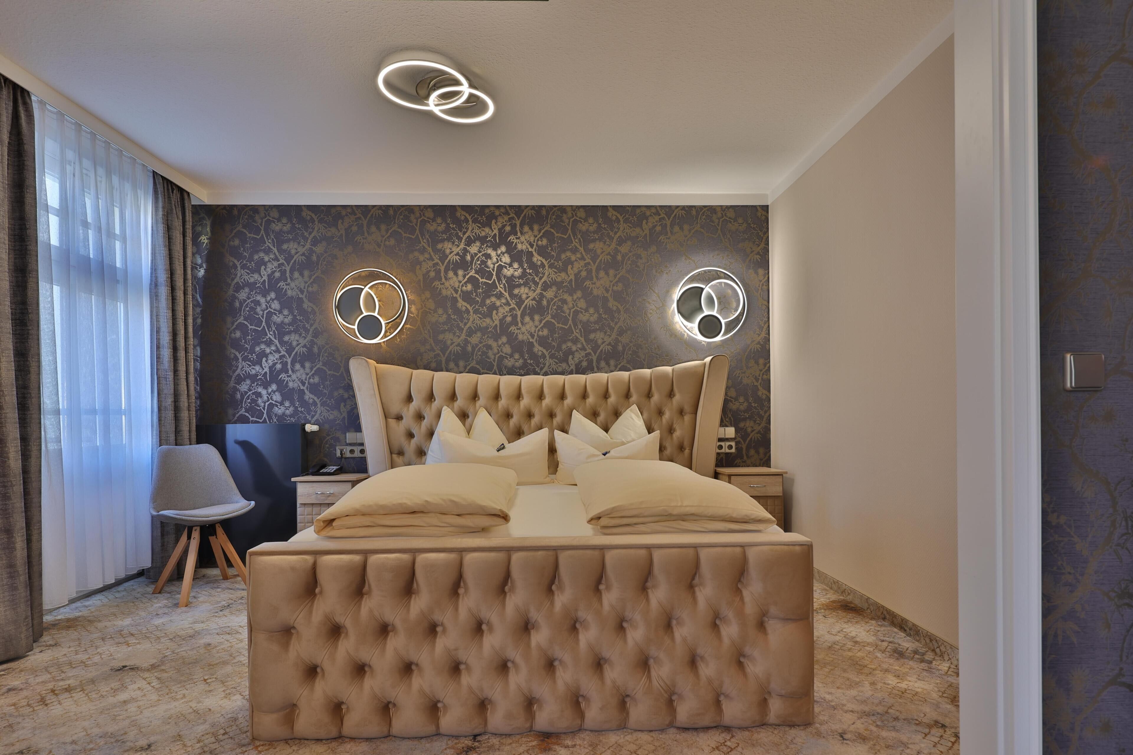 A modern bedroom with a tufted headboard, a large bed with multiple pillows, a bedside table, a chair, and a decorative wall with circular light fixtures.