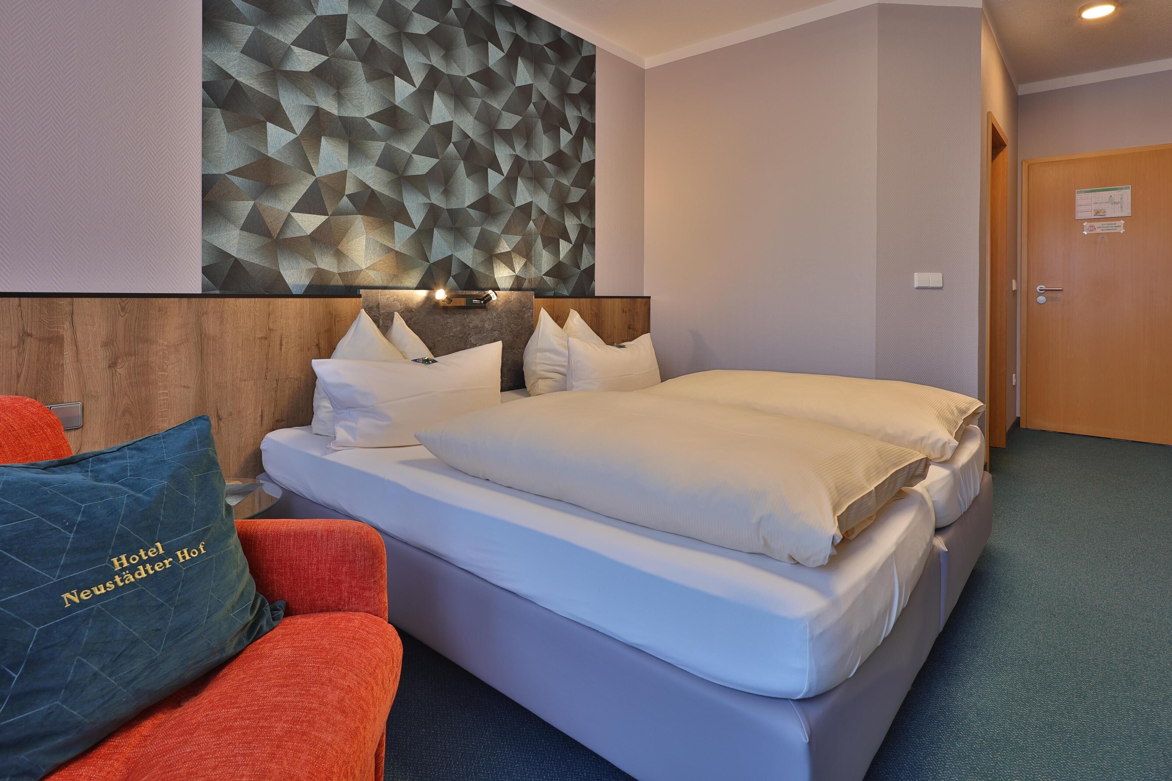 Habitación de hotel con cama, cabecera de madera, decoración mural geométrica, sillón rojo y almohada azul con texto.