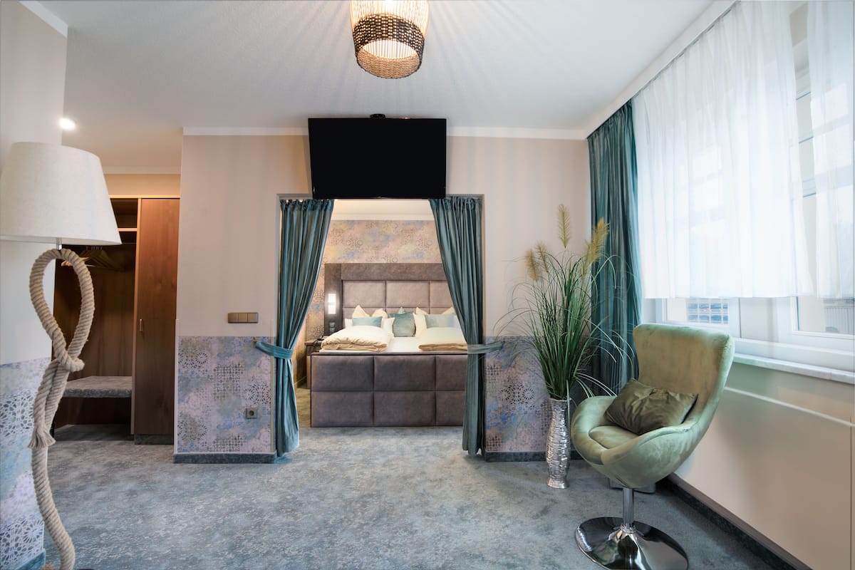A bedroom with a bed, a chair, a TV, and a window with curtains.