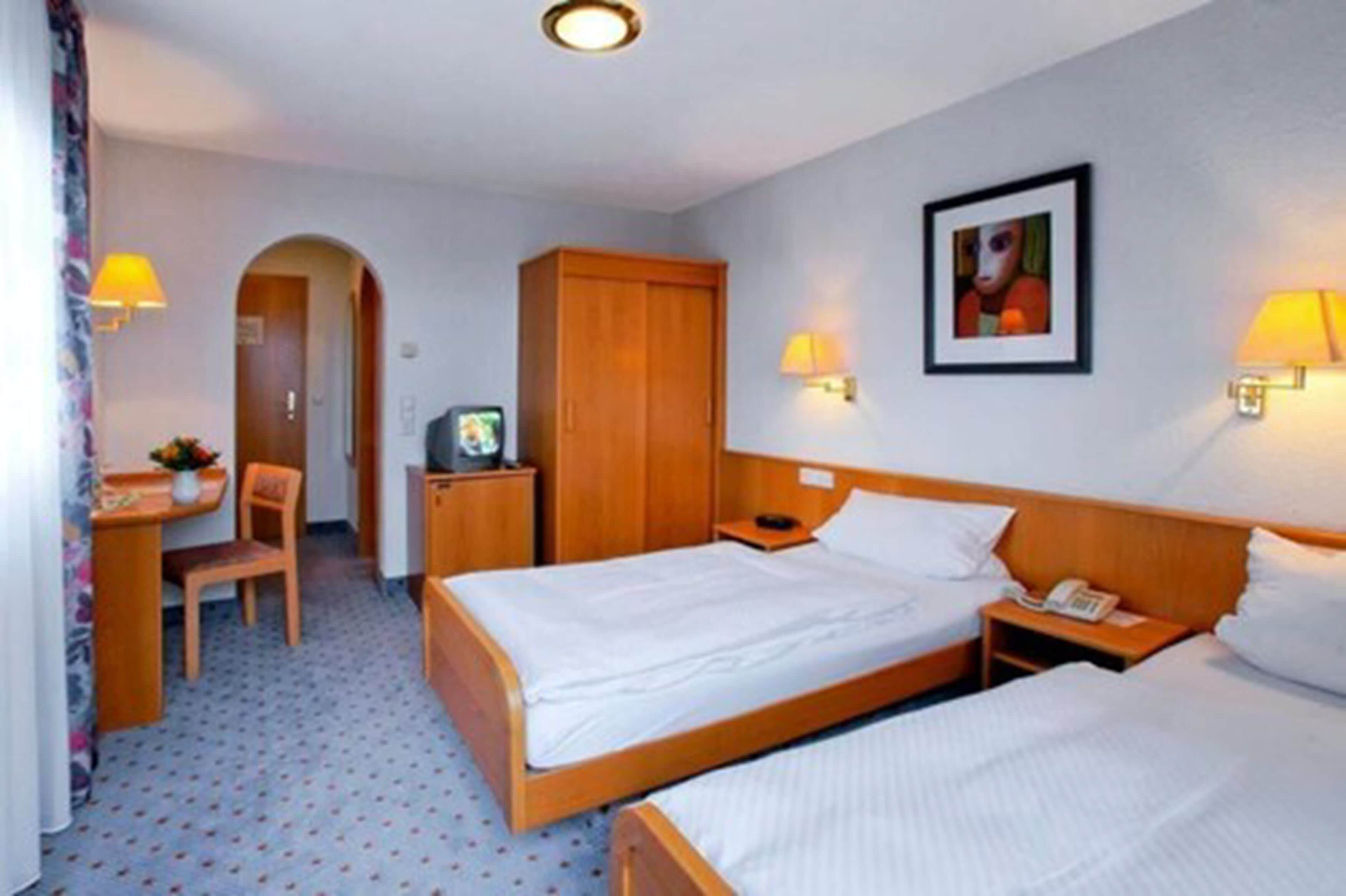 Habitación de hotel con dos camas, un escritorio, una silla, un armario y un cuadro en la pared.