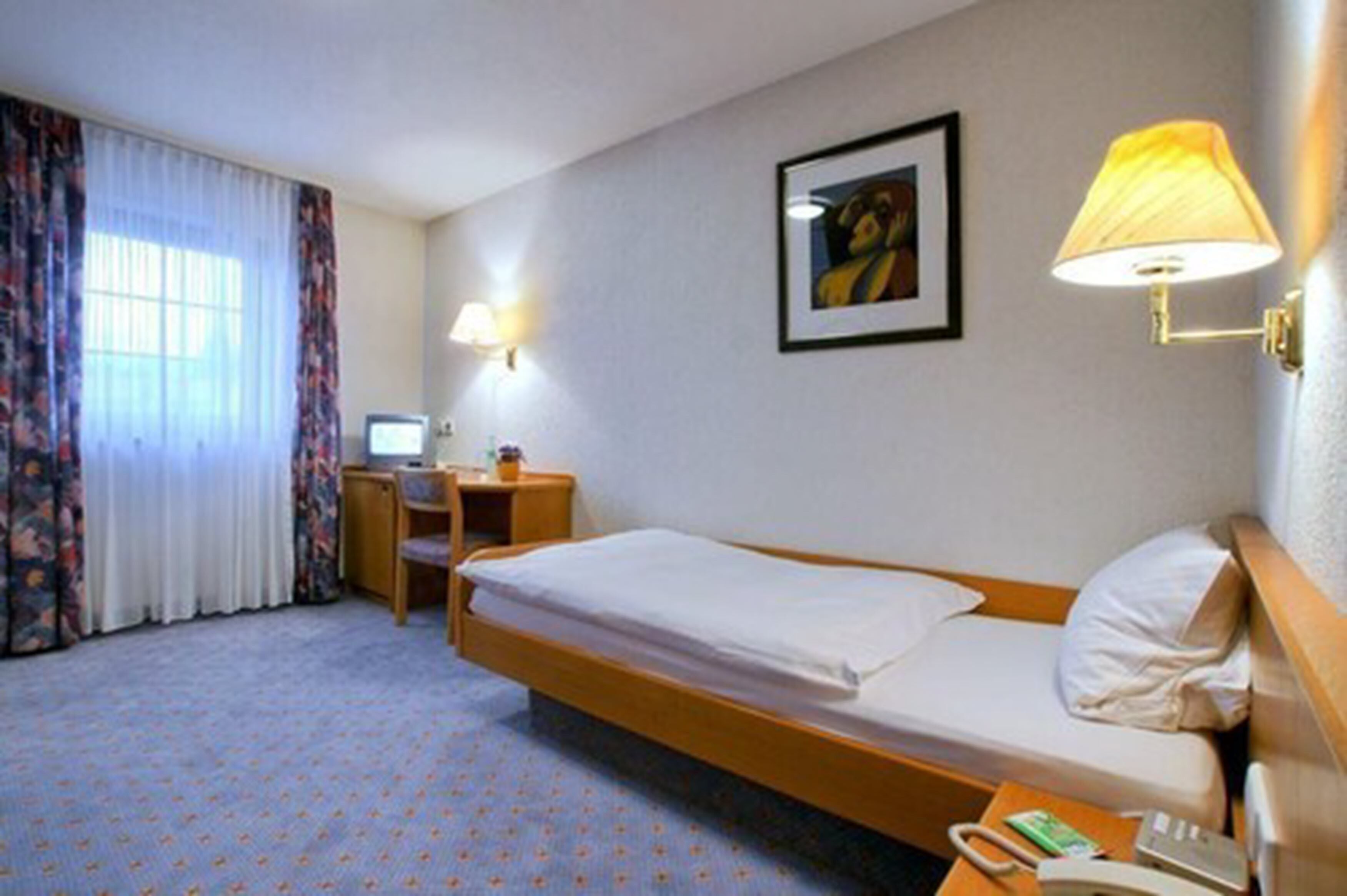 A hotel room with a single bed, a desk with a computer, a lamp, and a telephone.