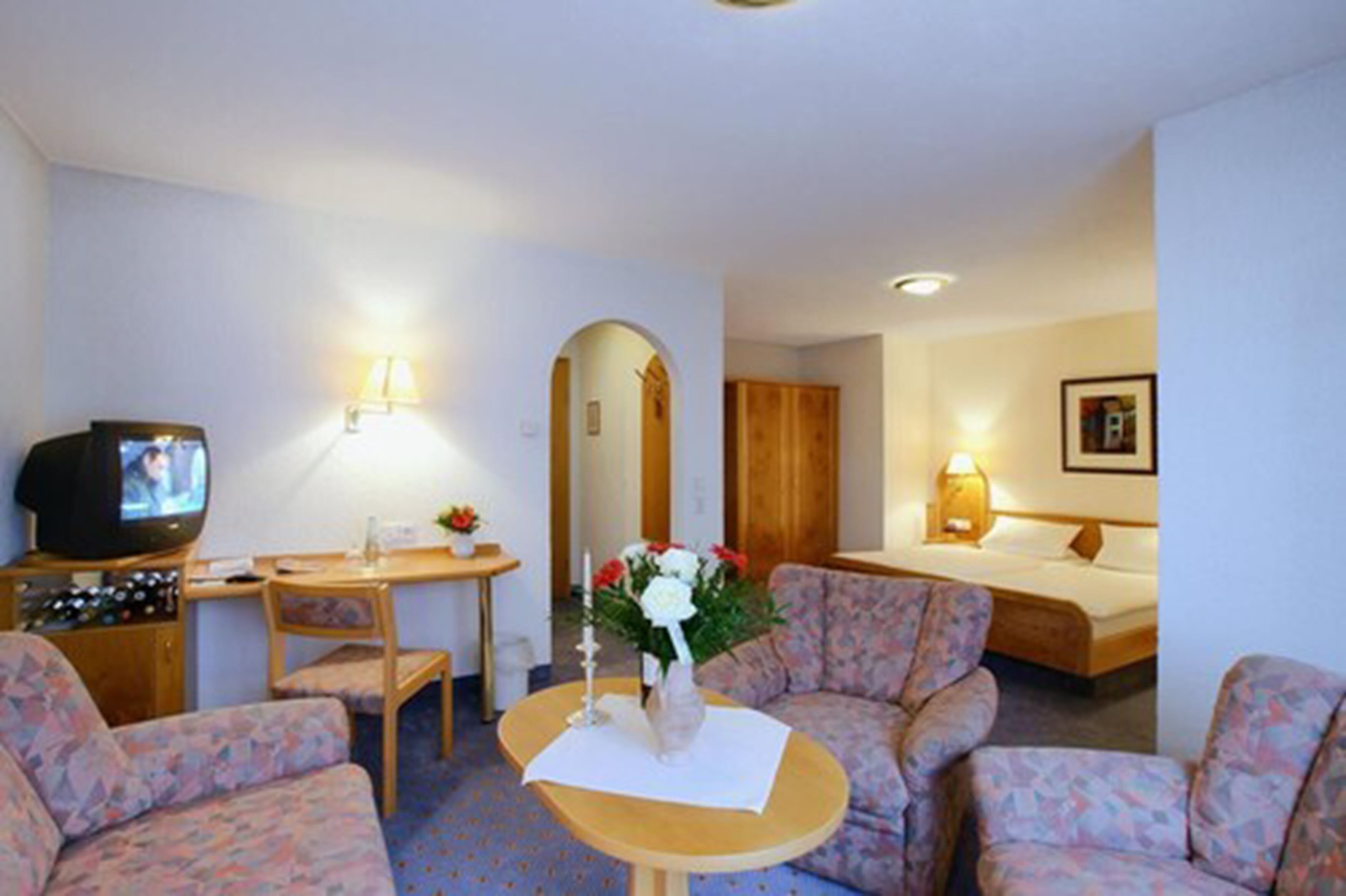 Une chambre d’hôtel avec un canapé, une petite table sur laquelle est posé un vase de fleurs, une télévision, un lit et un bureau avec une lampe.