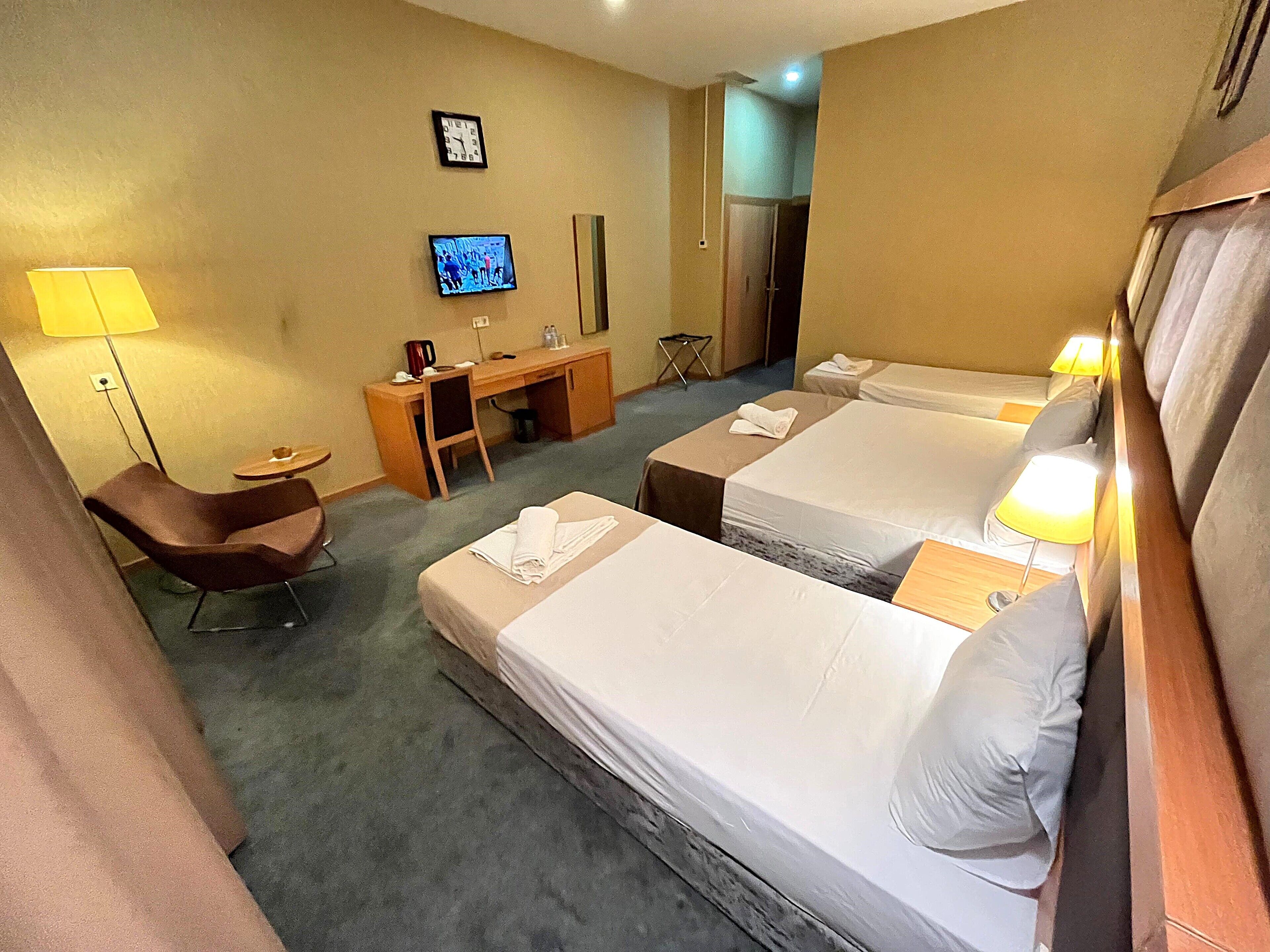 A hotel room with two beds, a desk, a chair, a TV, and a lamp.