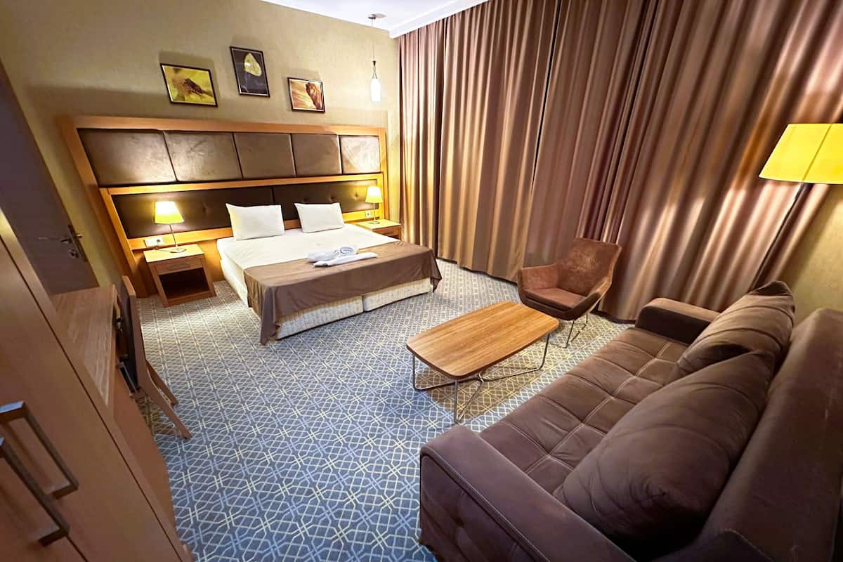 A hotel room with a large bed, a sofa, a small table, and a chair. There are curtains, a lamp, and a wall with pictures.