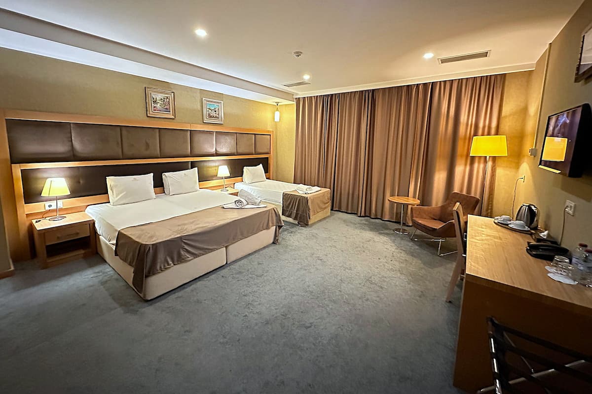 A hotel room with a large bed, two bedside tables with lamps, a desk with a chair, a TV, and a large window with curtains.