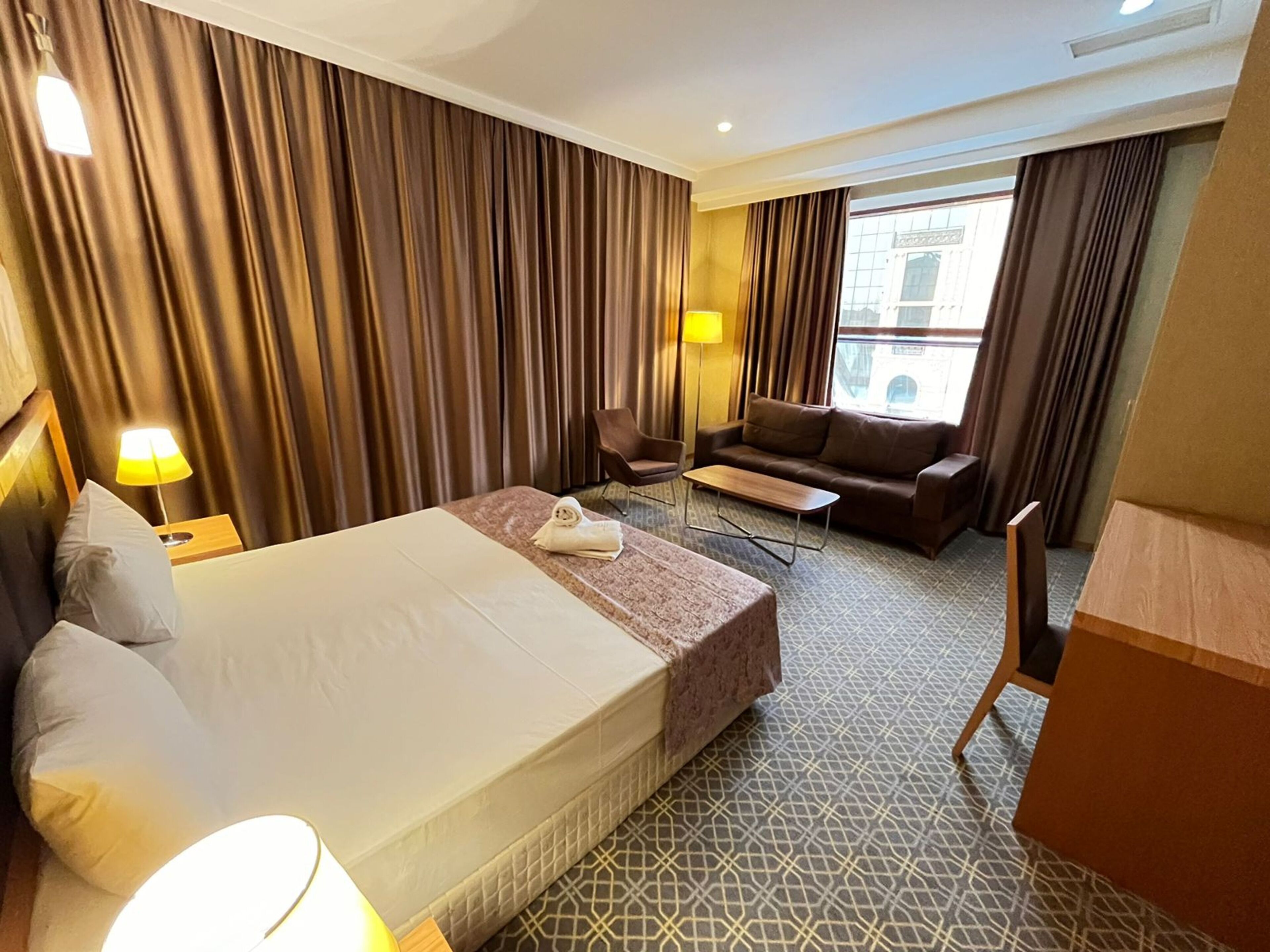 Habitación de hotel con una cama grande, un sofá, una mesita y una silla. Hay cortinas y una ventana con vista a un edificio.