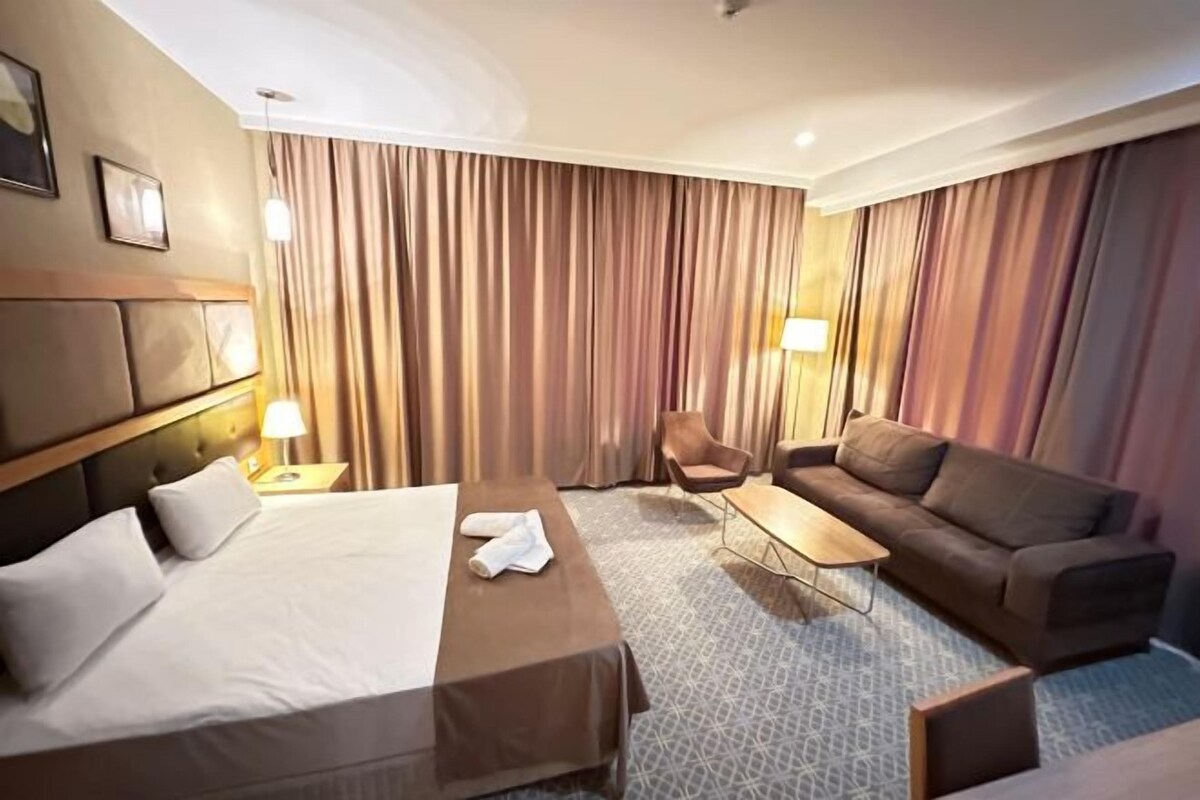 Quarto de hotel com cama, sofá, cadeira, mesinha e cortinas.
