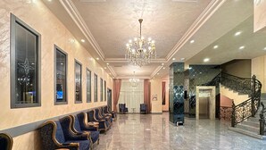 Un vestíbulo de hotel con una fila de sillones tapizados en azul, un candelabro y una escalera con barandales ornamentados.