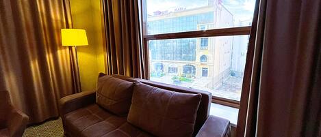 A hotel room with a brown leather sofa, a yellow lamp, and a view of a building through a large window.