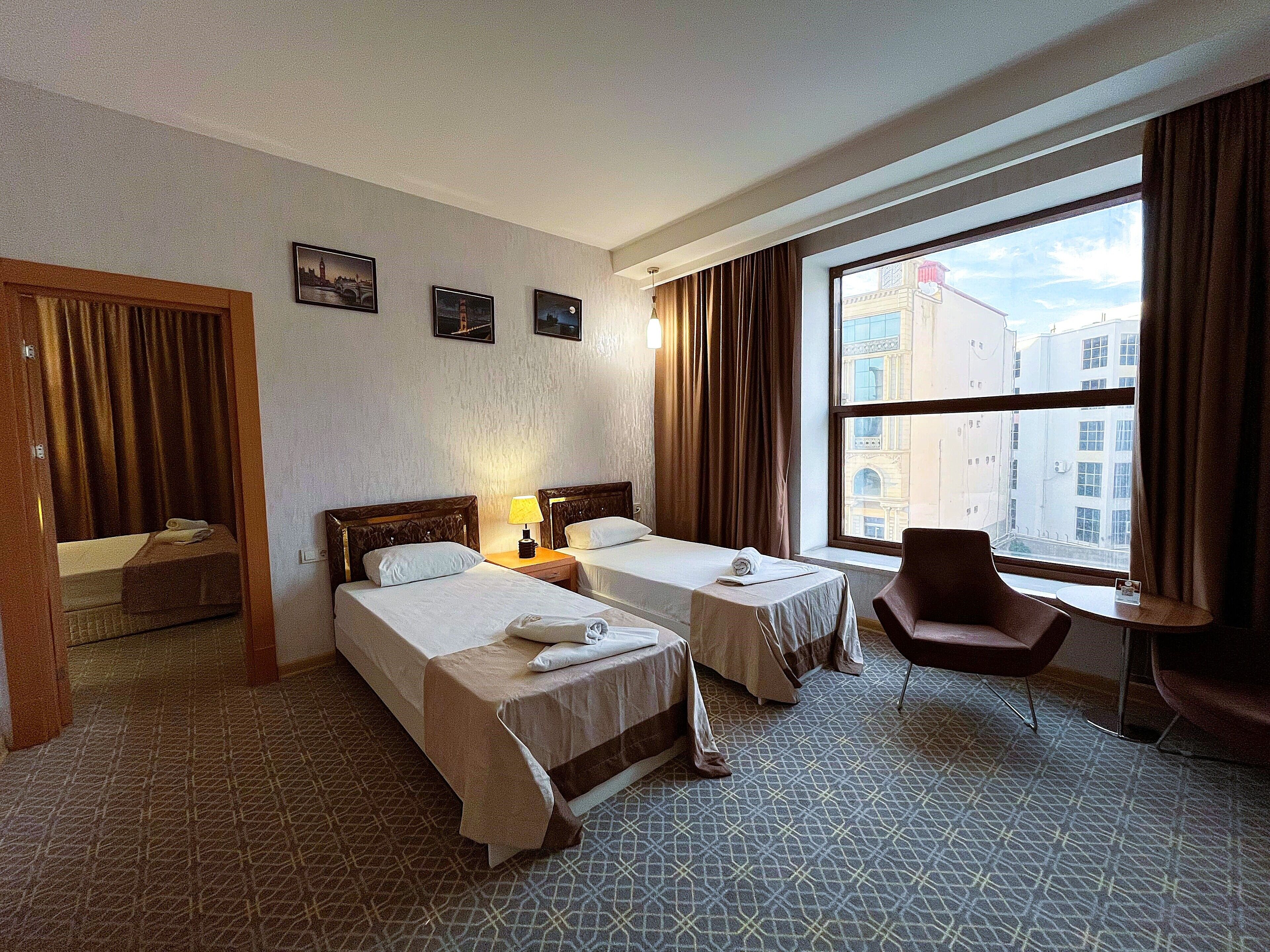 A hotel room with two beds, a chair, a desk, and a large window with a city view.
