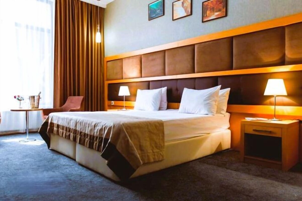 Ein Hotelzimmer mit einem großen Bett, zwei Nachttischen, einem Fenster mit Vorhängen und einer Wand mit gerahmten Bildern.