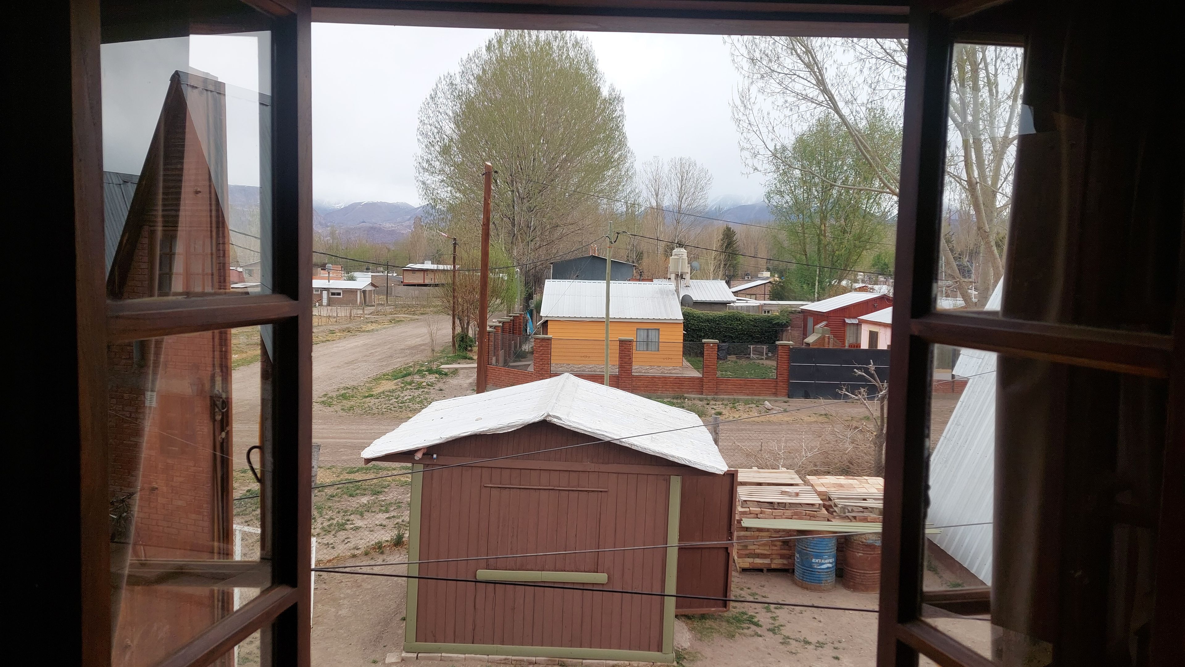 A view through a window of a rural area with a small wooden shed, a yellow house, and a dirt road.