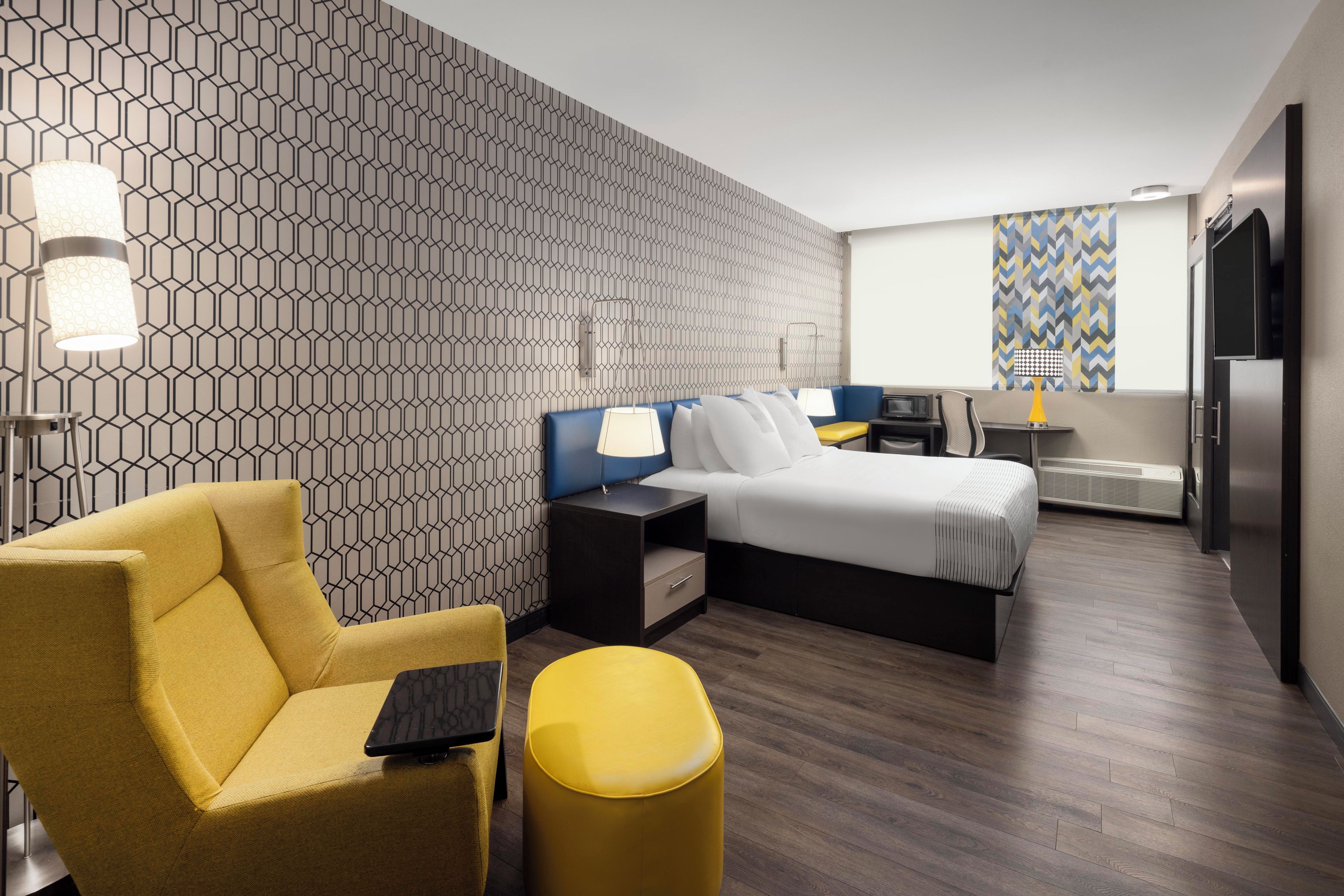 A modern hotel room with a geometric patterned wall, a bed, a desk, a chair, and a small ottoman.