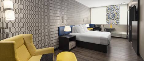 Moderni hotellihuone, jossa on geometrisesti kuvioitu seinä, sänky, työpöytä, tuoli ja keltainen rahi.