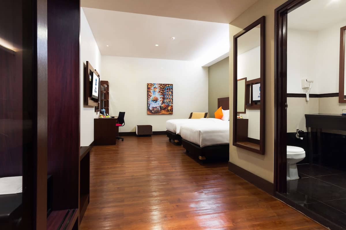 A hotel room with a wooden floor, a bed with white bedding, a painting on the wall, a desk with a chair, and a bathroom with a toilet and sink.