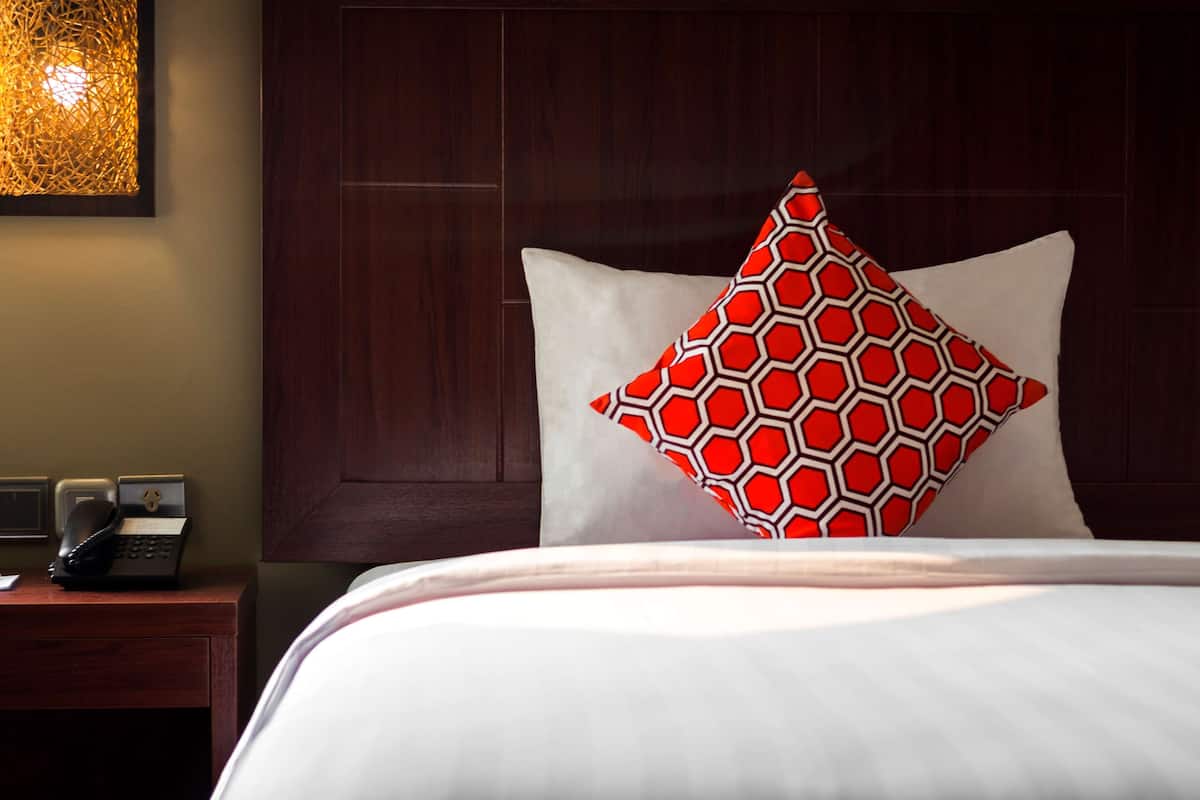 A hotel room with a neatly made bed, a wooden nightstand with a telephone, and a patterned pillow.
