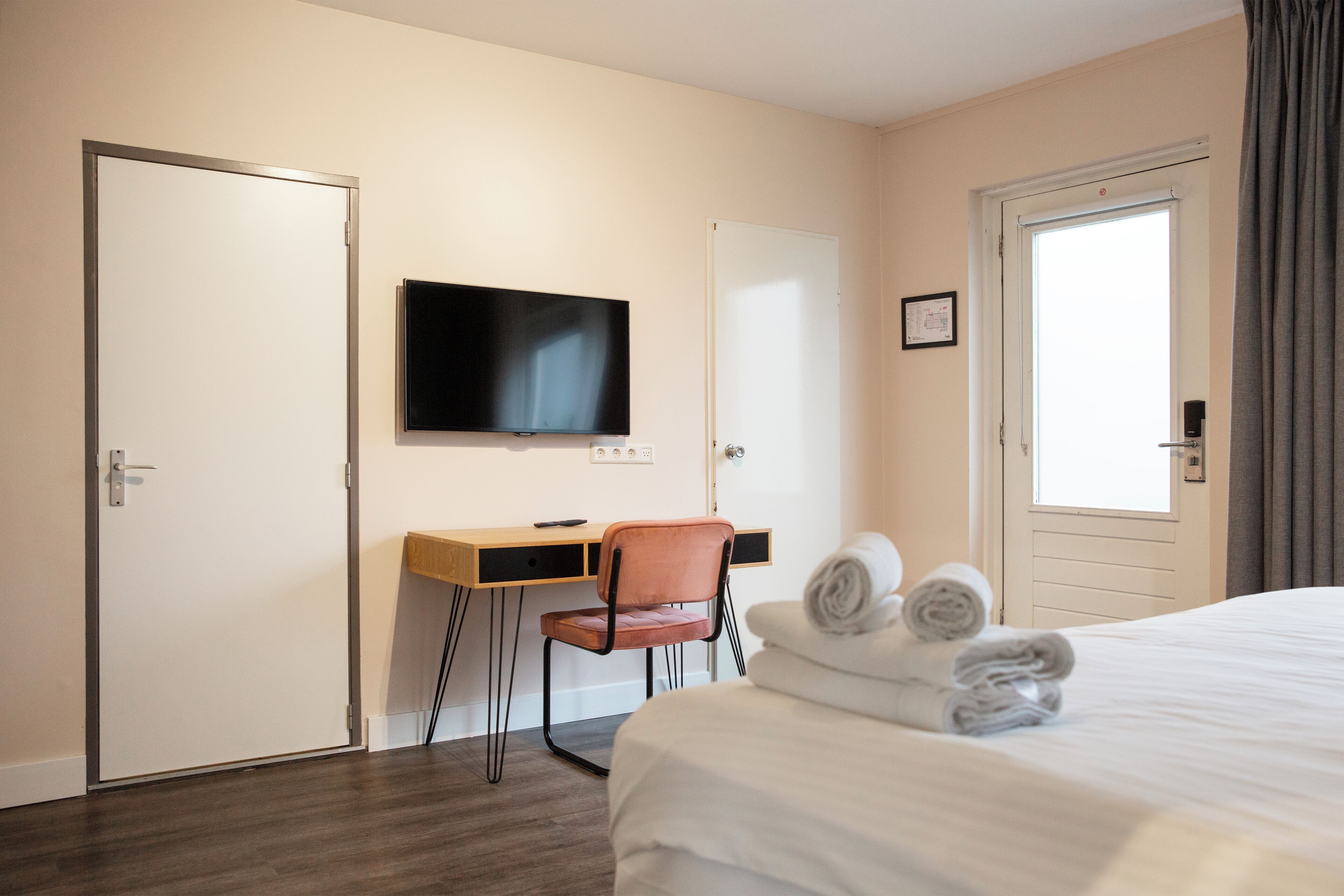 A hotel room with a bed, a desk with a chair, a television, and a door with a window.