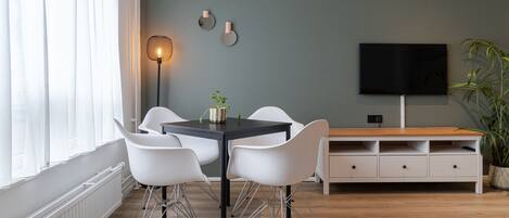 A modern dining area with a table and chairs, a floor lamp, a flat-screen TV, and a potted plant.