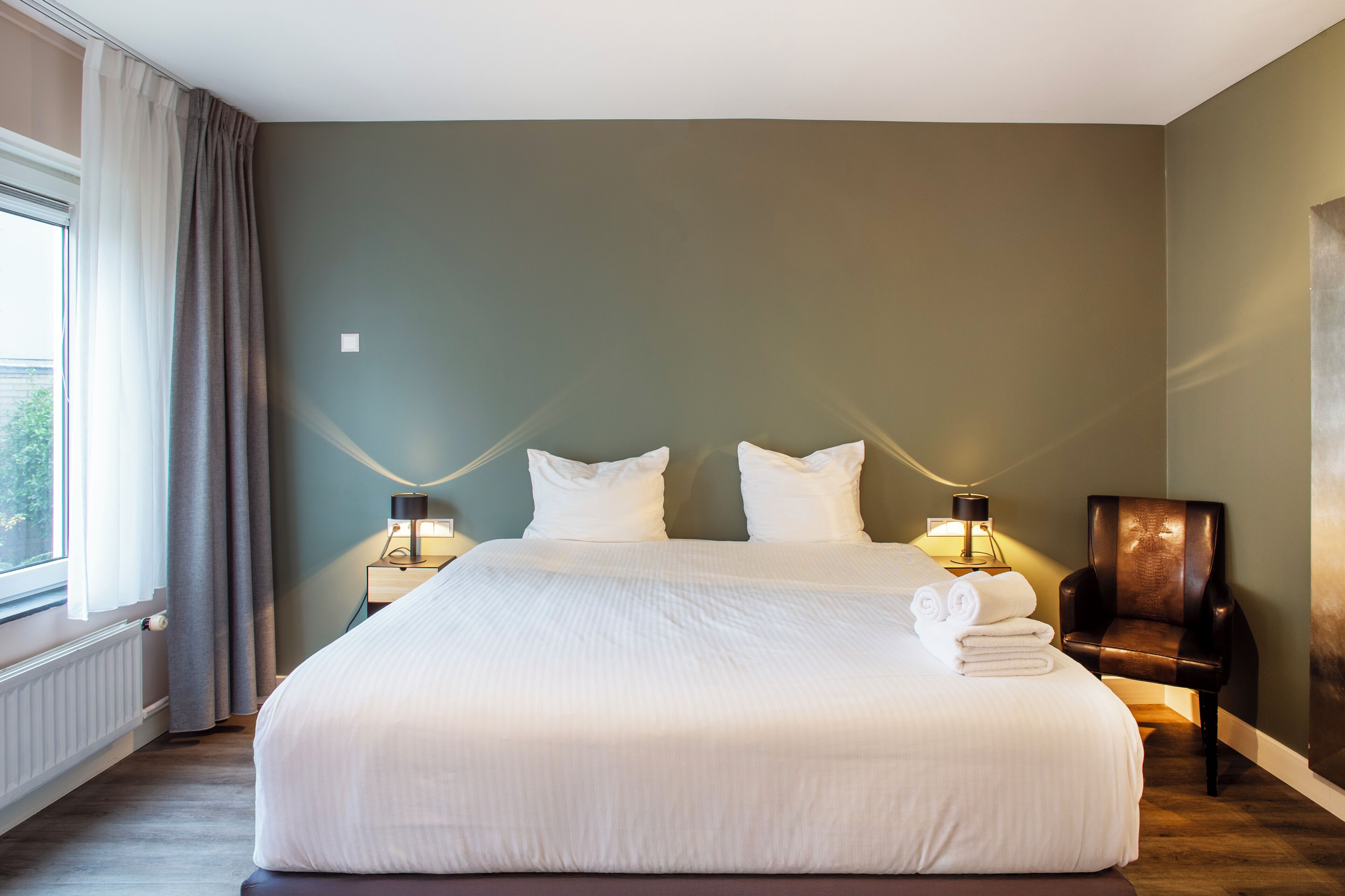 Una camera d'albergo con un letto grande, due lampade ai comodini, una poltrona e una finestra con tende.