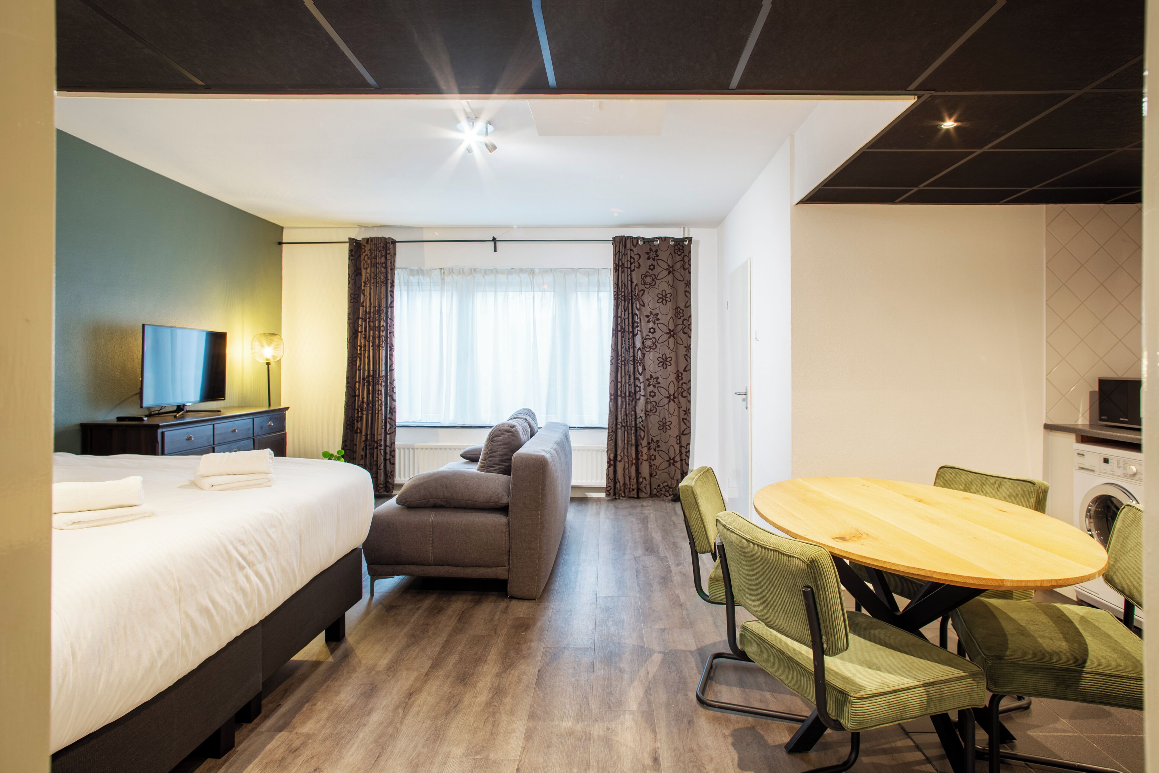 A hotel room with a bed, a sofa, a dining table with chairs, a television, and a kitchen area.