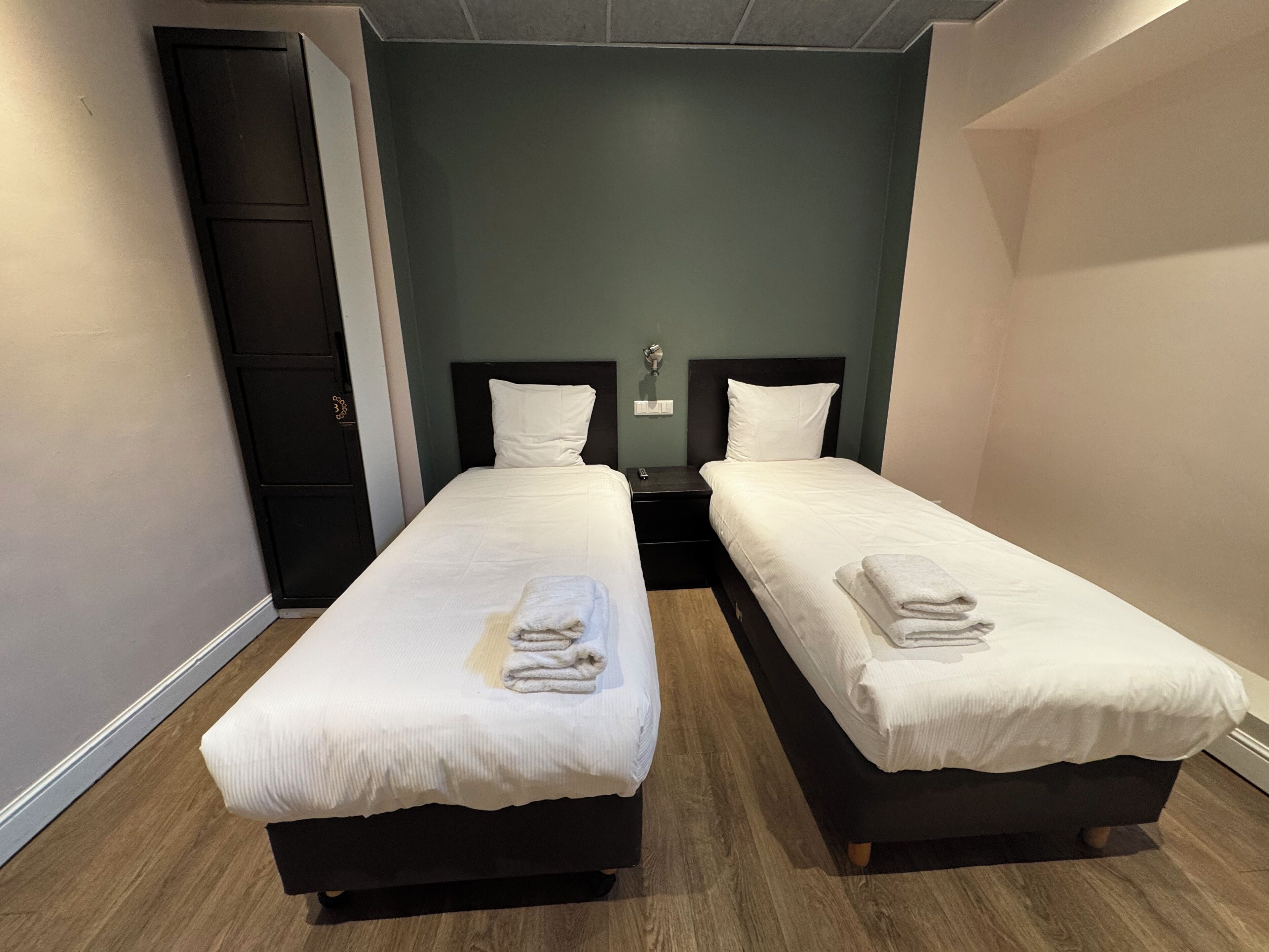 A room with two single beds, each with a white mattress and pillow, and a dark headboard. There is a wooden floor and a dark wardrobe to the left.