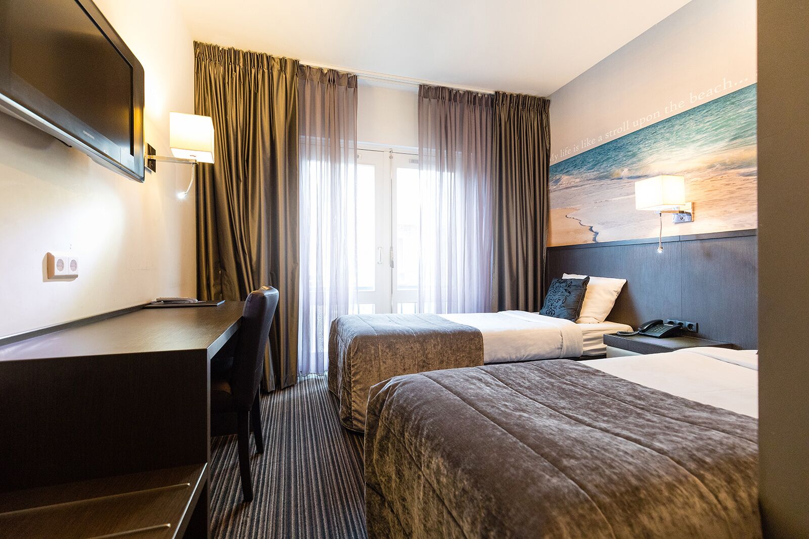 Habitación de hotel con escritorio, dos camas, un ventanal con cortinas y un cuadro en la pared.