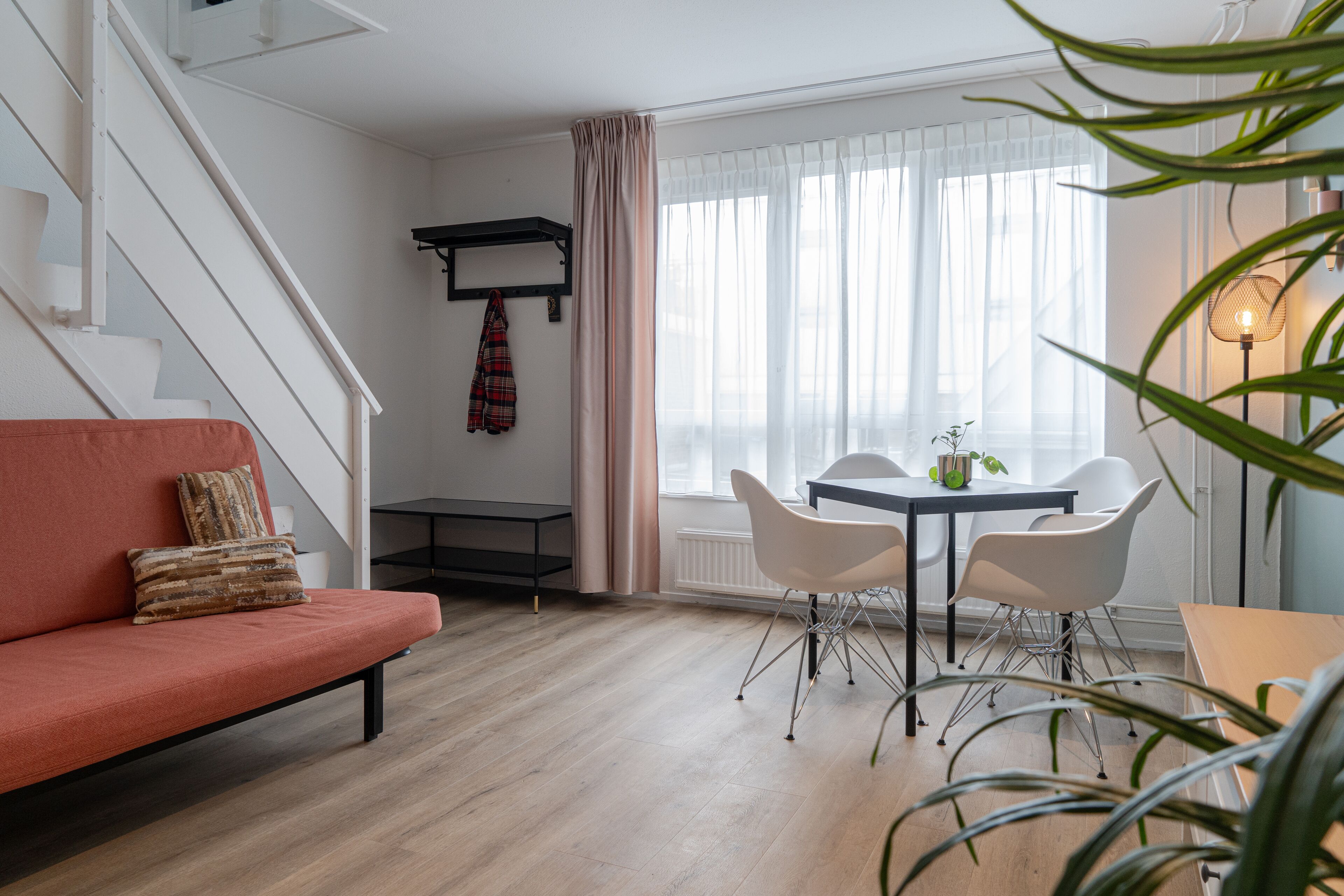 A modern living room with a sofa, a dining table with chairs, a staircase, and a window with curtains.