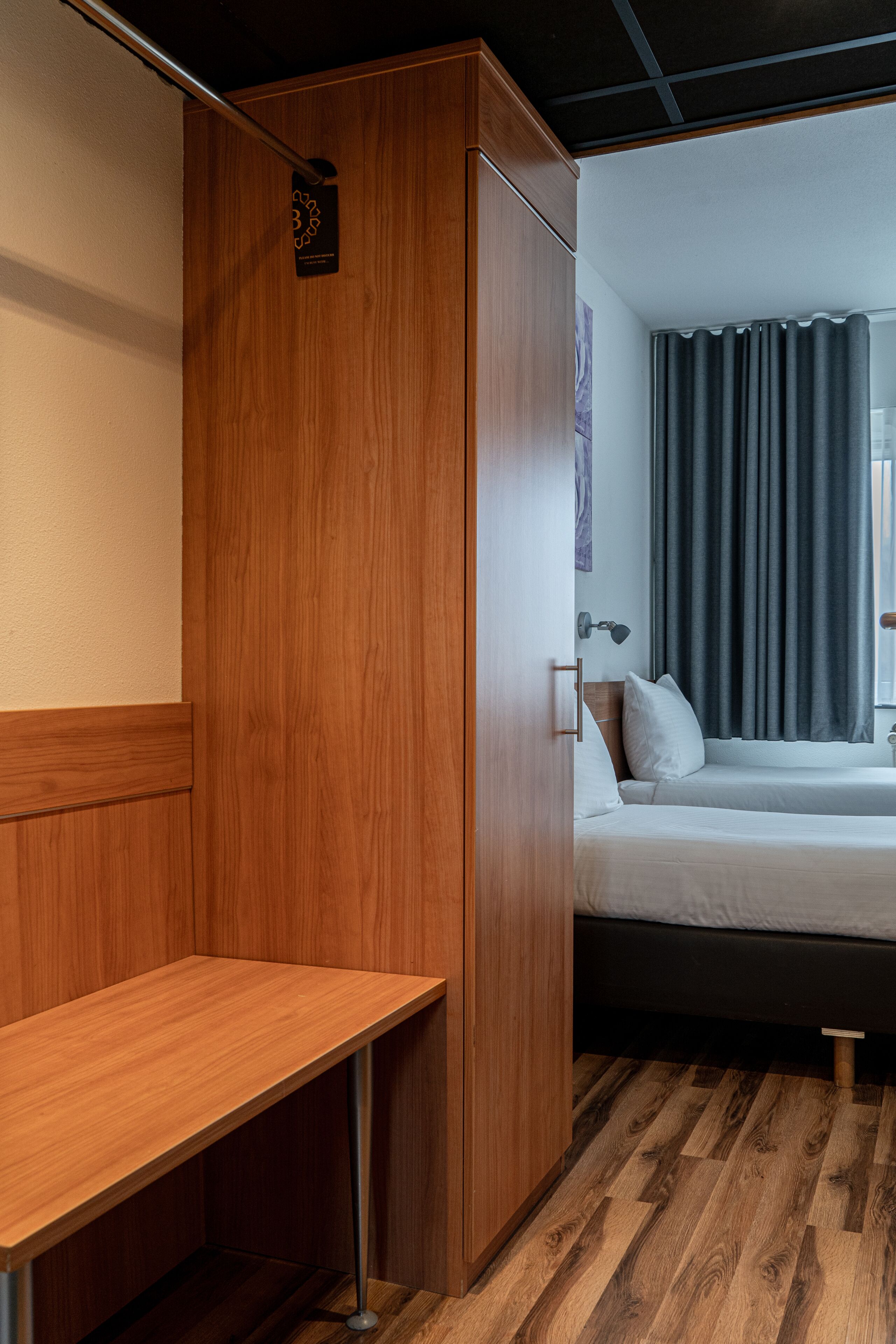 A hotel room with a wooden wardrobe, a bed with white bedding, a bedside table, and a window with curtains.