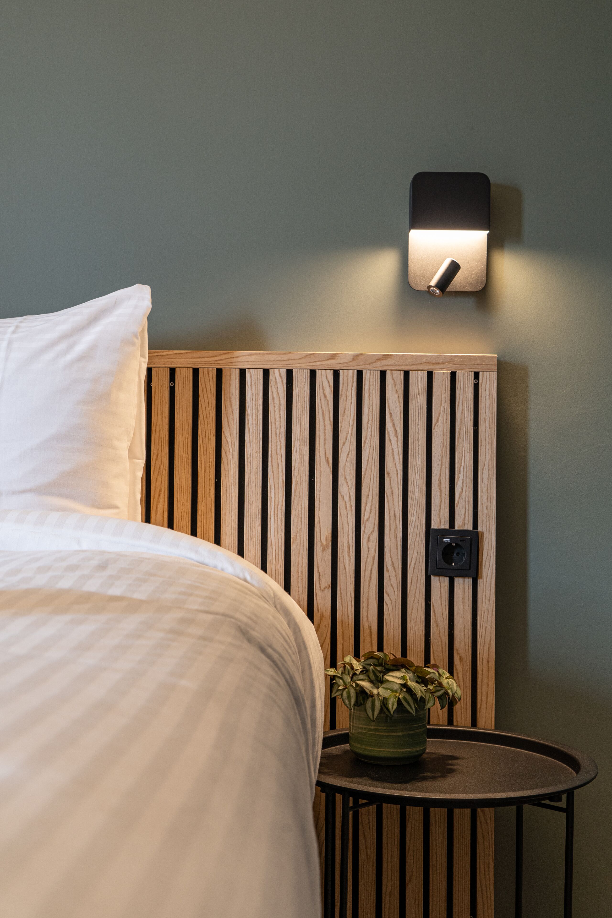 A neatly made bed with white linens, a wooden slatted headboard, a small round table with a potted plant, and a wall-mounted light fixture.
