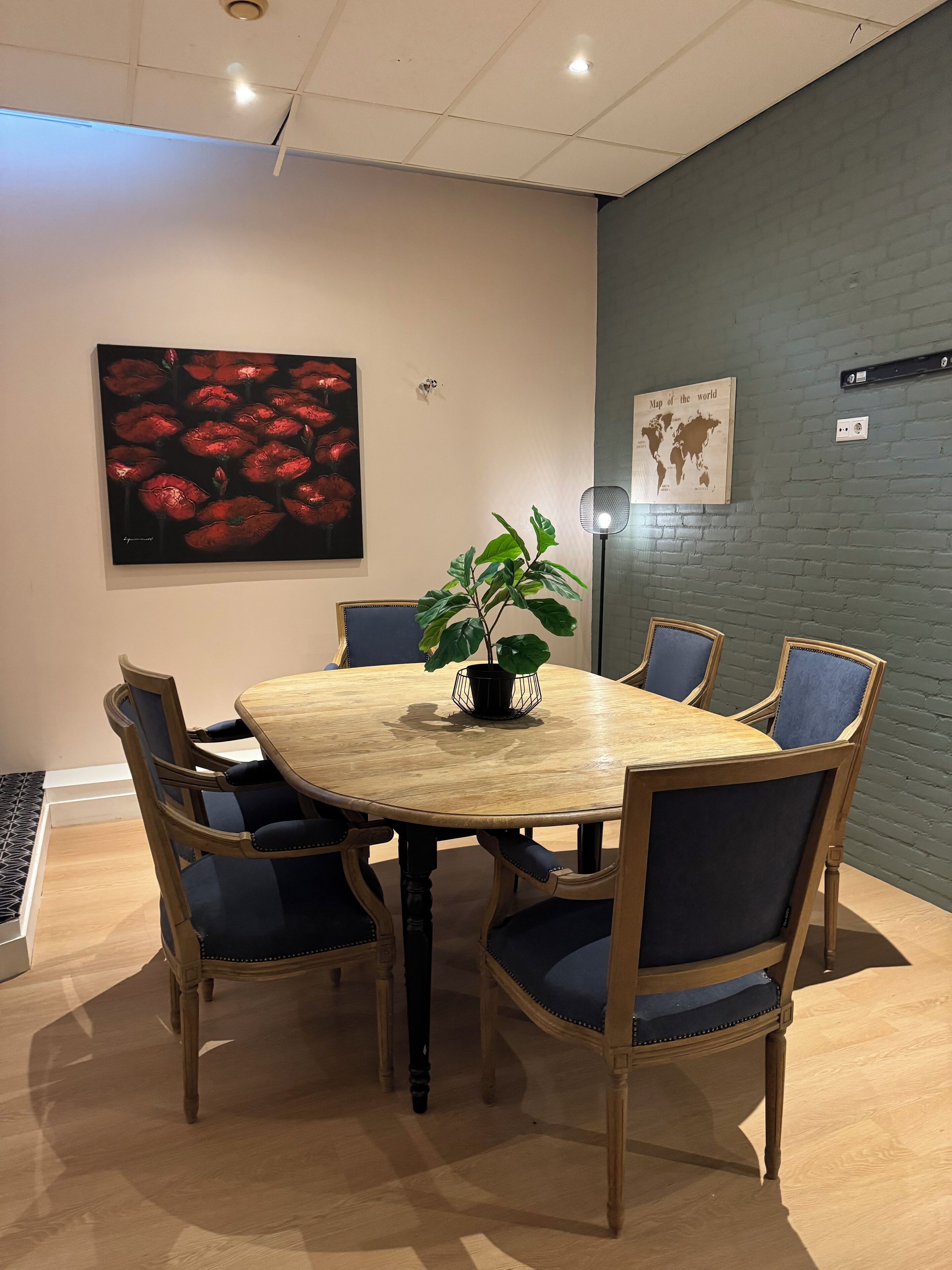 A conference room with a round wooden table, six chairs, a potted plant, a world map on the wall, and a painting of red flowers.