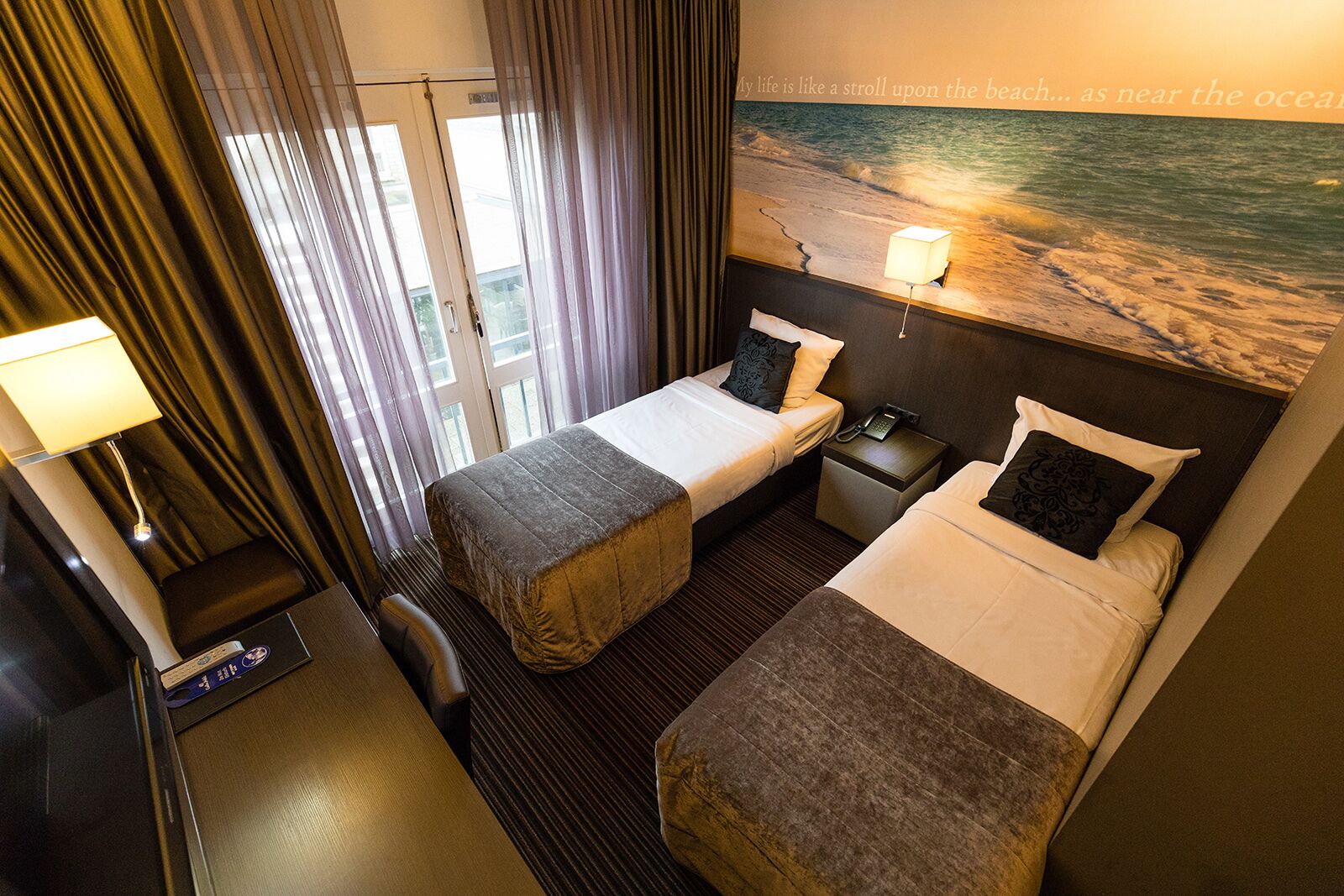 A hotel room with two beds, a desk, a chair, a lamp, and a large window with curtains.