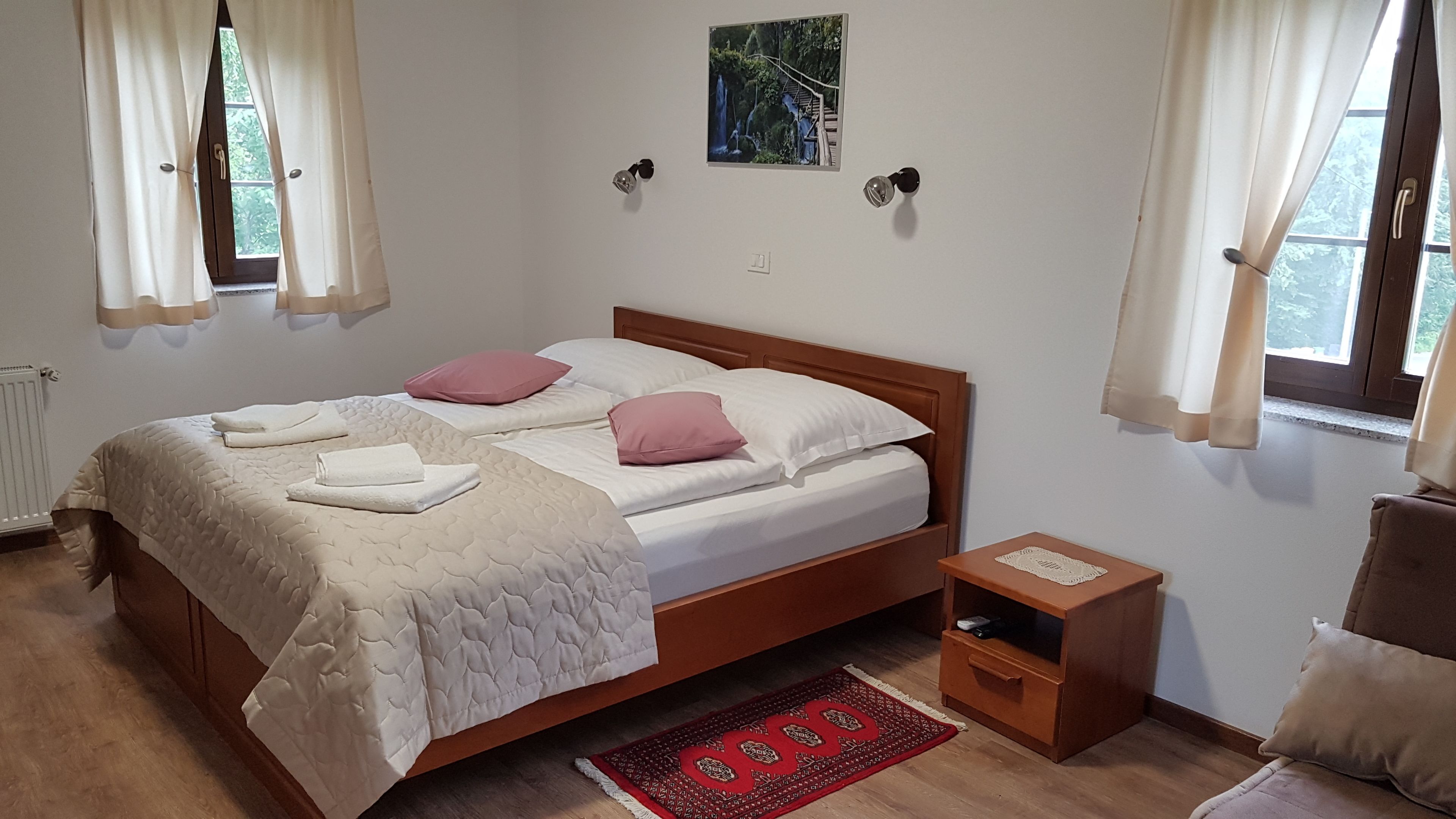 Un dormitorio con una cama, dos almohadas, una mesita de noche, una pequeña mesa, una alfombra roja y un cuadro en la pared.