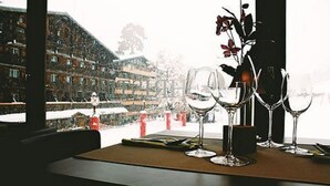 Uma mesa com copos de vinho e um vaso de flores, com vista para uma paisagem nevada com edifícios.