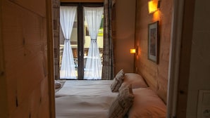 Corredor estreito que dá para uma cama com lençóis brancos e almofadas estampadas. Um porta de madeira e uma luminária na parede são visíveis.