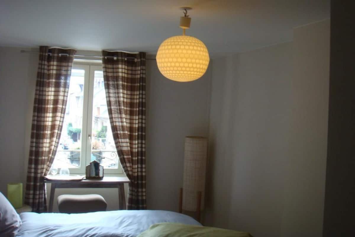 Een slaapkamer met een bed, een raam met gordijnen, een tafel met lamp en een hanglamp.