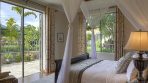 Un dormitorio con una cama grande, una mesita de noche con lámpara, vistas al jardín a través de la ventana y un balcón con plantas en macetas.