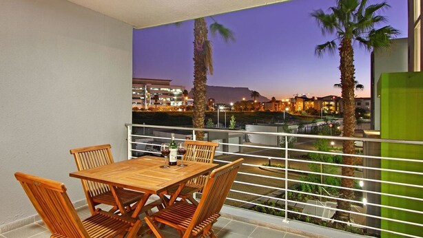 Ein Balkon mit Holztisch und -stühlen, der auf eine städtische Szenerie im Abendlicht mit Palmen und Gebäuden im Hintergrund blickt.