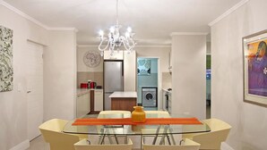 Ein moderner Essbereich mit einem Glastisch, vier Stühlen, einer Pendelleuchte und einer Küche im Hintergrund.