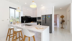 Uma cozinha moderna com armários brancos, uma ilha central com banquetas, eletrodomésticos de aço inoxidável e iluminação pendente.