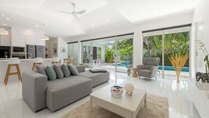 Uma sala de estar moderna com um sofá seccional cinza, uma mesa de centro branca e uma poltrona cinza. Há uma porta de vidro que dá para uma área de piscina com vista para o verde.