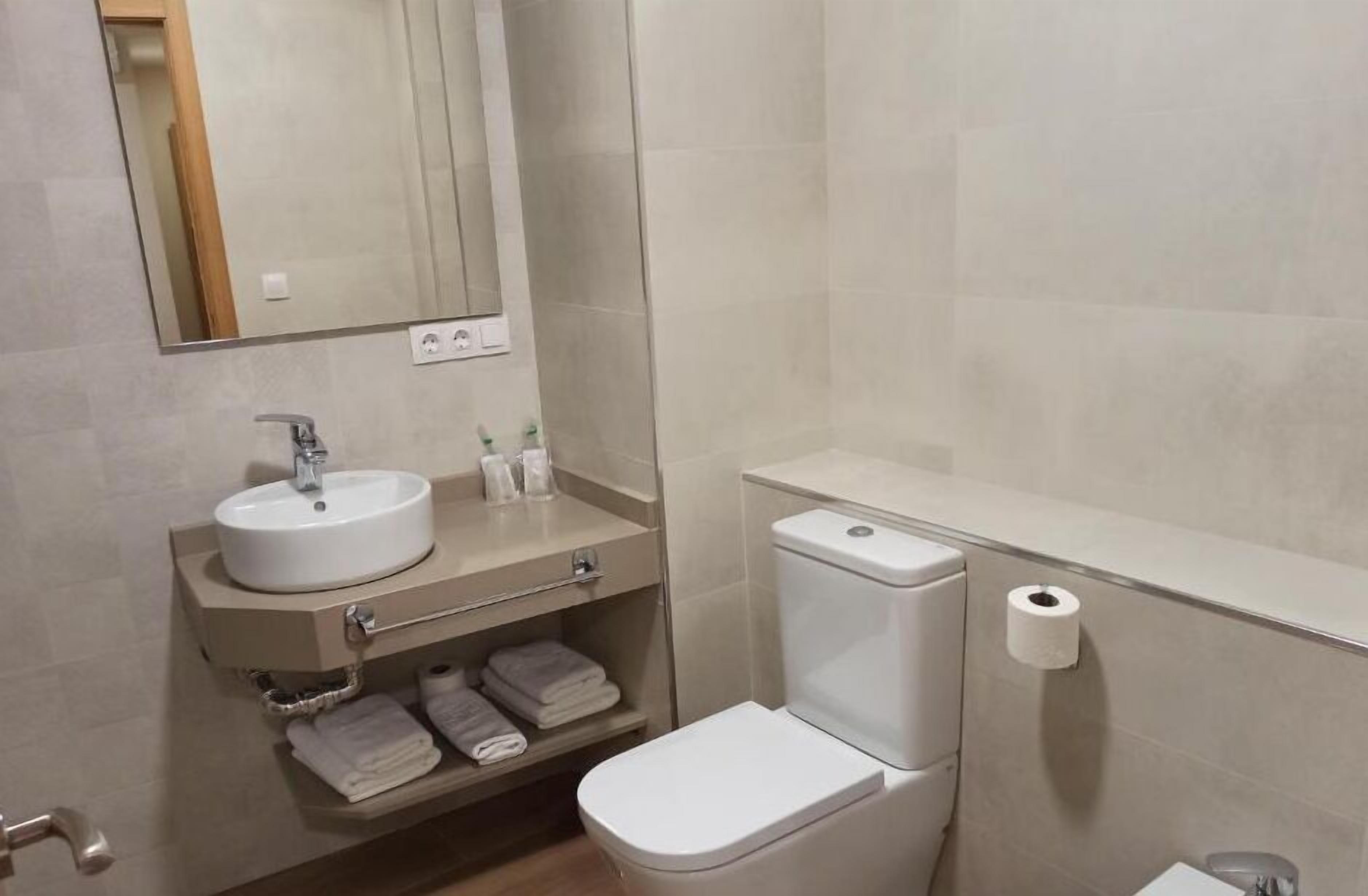 A bathroom with a white sink, mirror, and toilet. There are towels on the shelf and a toilet paper roll on the wall.
