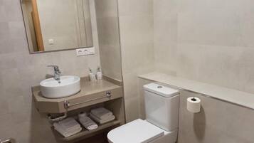 A bathroom with a white sink, mirror, and toilet. There are towels on the shelf and a toilet paper roll on the wall.