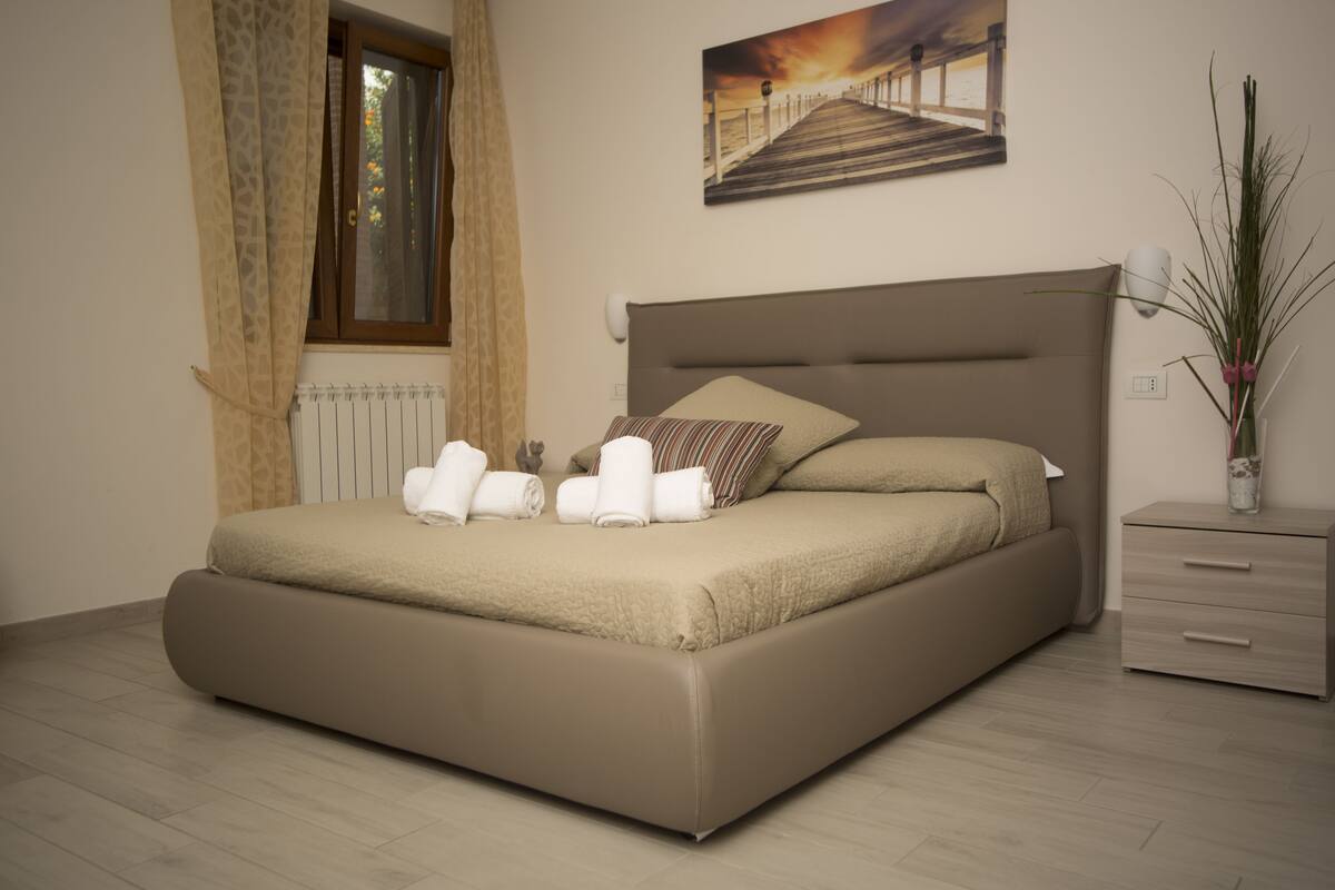 A bedroom with a large bed, a nightstand, a window with curtains, a painting on the wall, and a potted plant.