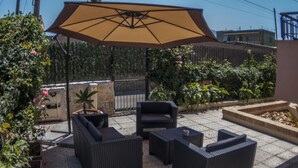 Uma área de terraço ao ar livre com móveis de vime, um grande guarda-sol e plantas em vasos.