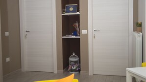 Quarto com duas portas brancas, uma mesa branca e uma cadeira amarela. Há uma estante com livros e uma gaiola de pássaros.