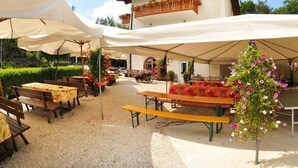 An outdoor seating area with wooden benches and tables, covered by white canopies, and surrounded by greenery. - Albergo Nido Verde (Lavarone)