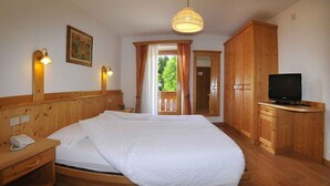 A wooden-furnished bedroom with a bed, bedside table with a phone, a television, and a large window with curtains. - Albergo Nido Verde (Lavarone)