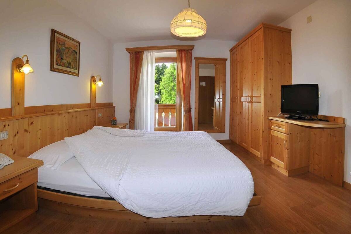 A wooden-furnished bedroom with a bed, bedside table with a phone, a television, and a large window with curtains.