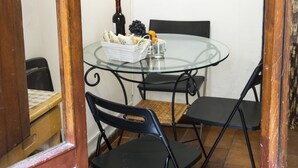 Uma pequena área de refeições com mesa de vidro, cadeiras pretas, uma cesta branca com pão e uvas, e uma garrafa de vinho.